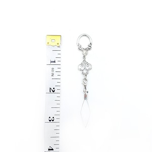Pode incluir: Brinco prateado com um design ornamentado em filigrana e um pingente pendurado em forma de diamante transparente. O brinco tem aproximadamente 7,6 cm de comprimento, medido com uma fita m&eacute;trica amarela e branca.