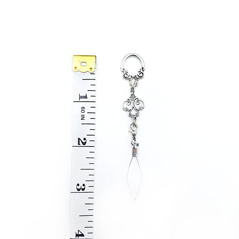 Pode incluir: Brinco prateado com um design ornamentado em filigrana e um pingente pendurado em forma de diamante transparente. O brinco tem aproximadamente 7,6 cm de comprimento, medido com uma fita m&eacute;trica amarela e branca.