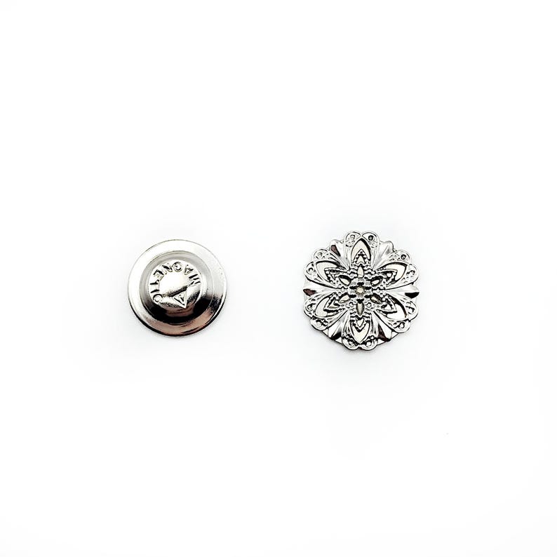 May include: A silver-toned metal button with a decorative floral design. The button has a flat back with a small pin.