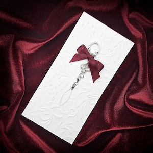 Pode incluir: Um chaveiro decorativo com um la&ccedil;o bord&ocirc; e um pingente transparente em forma de diamante. O chaveiro est&aacute; preso a um cart&atilde;o branco em relevo com um padr&atilde;o floral, sobre um fundo de veludo vermelho escuro.