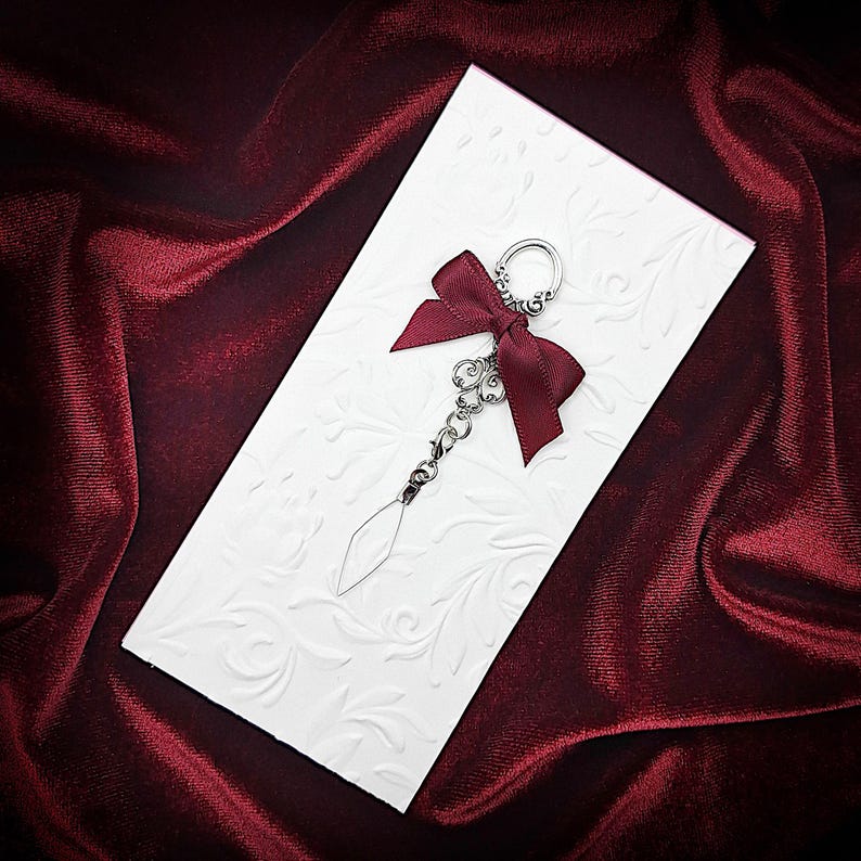 Pode incluir: Um chaveiro decorativo com um la&ccedil;o bord&ocirc; e um pingente transparente em forma de diamante. O chaveiro est&aacute; preso a um cart&atilde;o branco em relevo com um padr&atilde;o floral, sobre um fundo de veludo vermelho escuro.