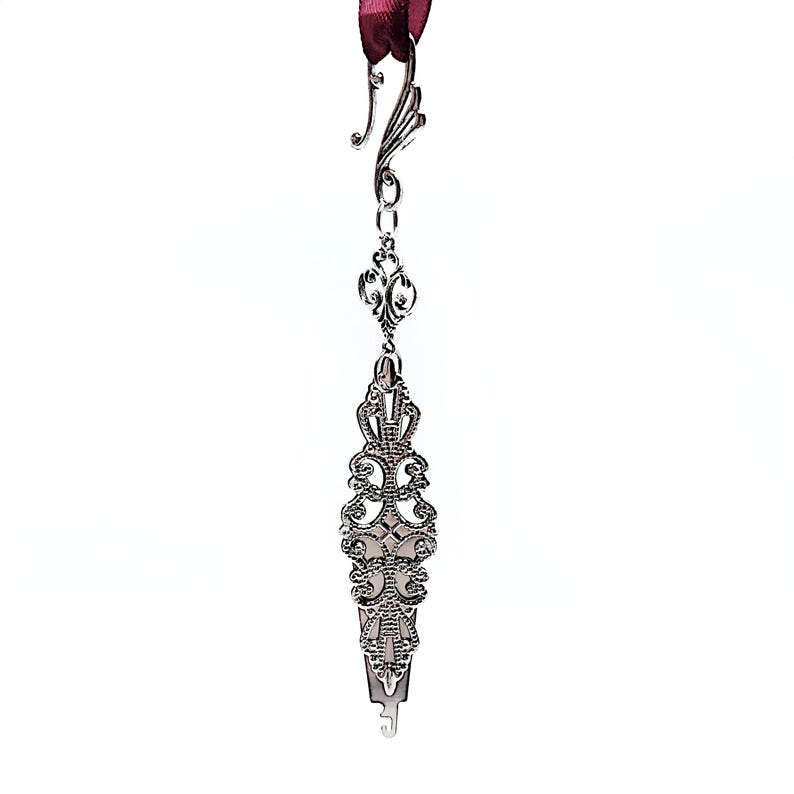 May include: Silver bookmark with an ornate design and a hook at the top. The bookmark is attached to a red ribbon.