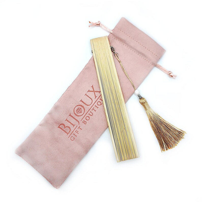 May include: A gold-colored folding fan with a tassel and a pink velvet pouch. The pouch has the text "Bijoux Gift Boutique" printed on it.
