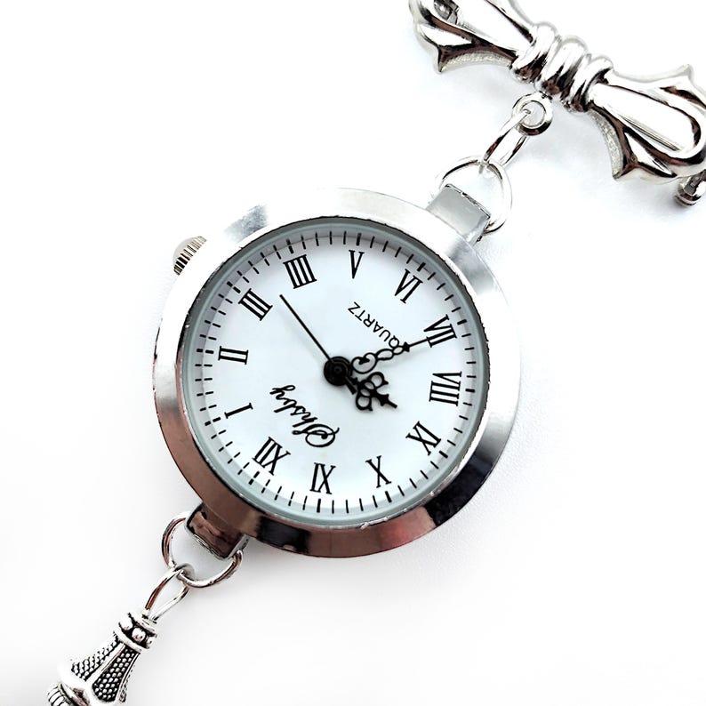 May include: A silver pocket watch with a white face and black Roman numeral hour markers. The watch has a chain attached to a decorative silver pendant.
