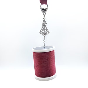 May include: A spool of red thread with a white base is suspended by a decorative silver charm. The charm features an ornate design with a butterfly at the top and a fleur-de-lis shape below. A maroon ribbon is attached to the top.