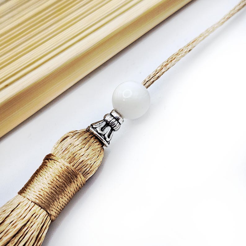 May include: A beige tassel with a silver metal cap and a white bead on a beige cord.