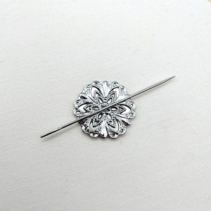 May include: A silver metal decorative piece with a floral design, pierced with a silver needle.