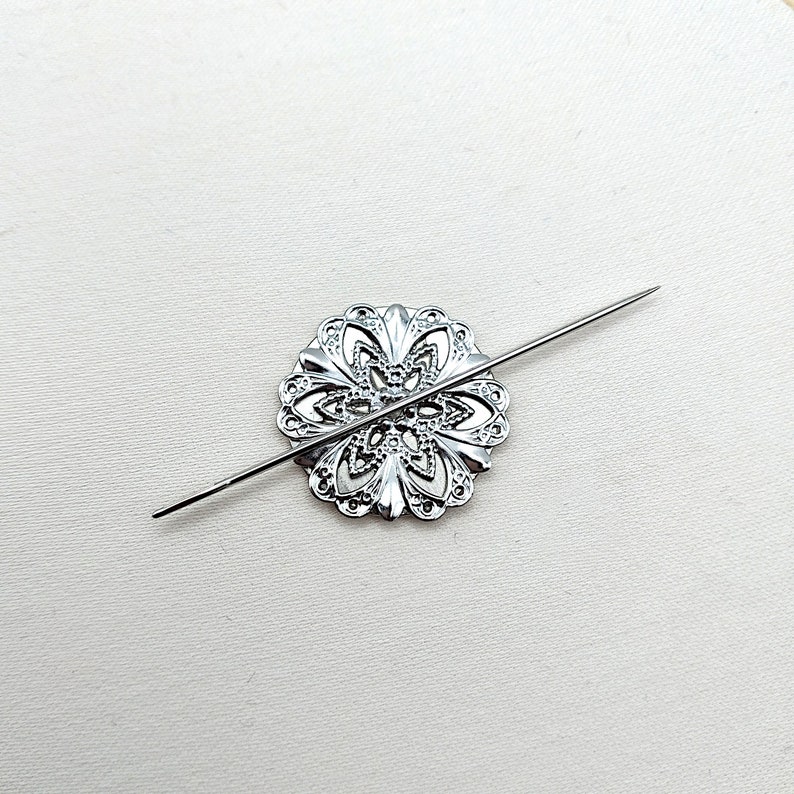 May include: A silver metal decorative piece with a floral design, pierced with a silver needle.