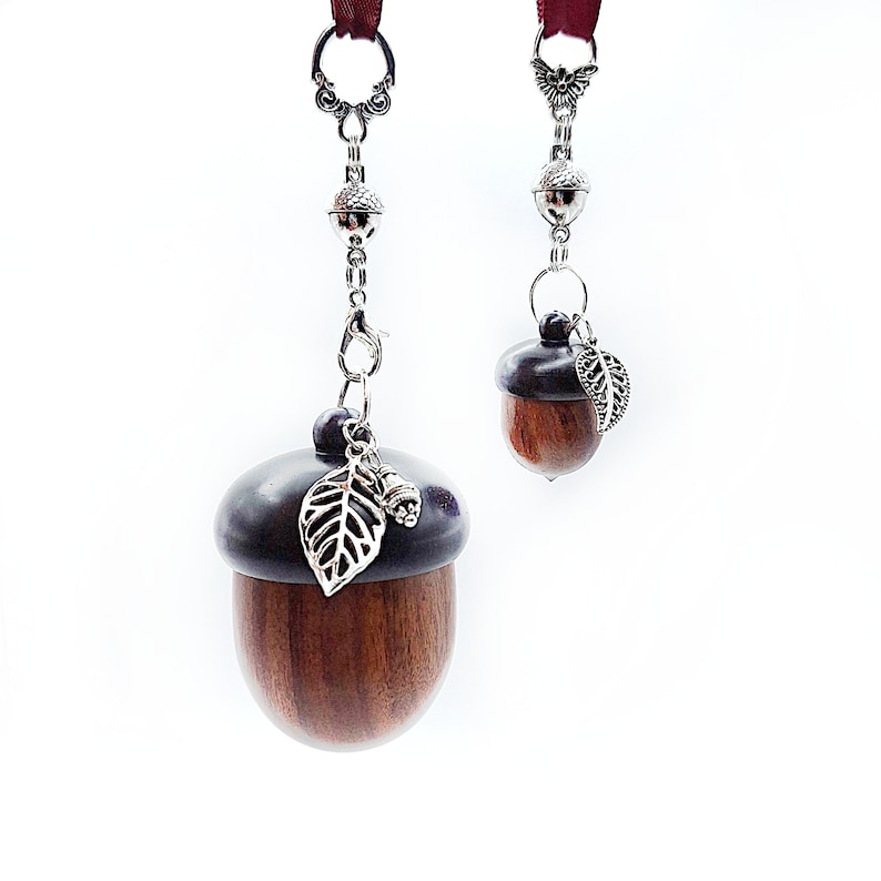 May include: Two wooden acorn-shaped charms with silver chains and leaf accents. The larger charm has a dark brown finish and the smaller charm has a lighter brown finish.