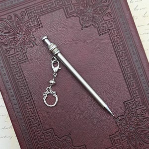 May include: A silver-toned metal mechanical pencil featuring a slender, smooth barrel and a decorative cap with a ribbed texture. A small silver charm in the shape of an ornate oval frame is attached to the pencil by a lobster clasp and chain.