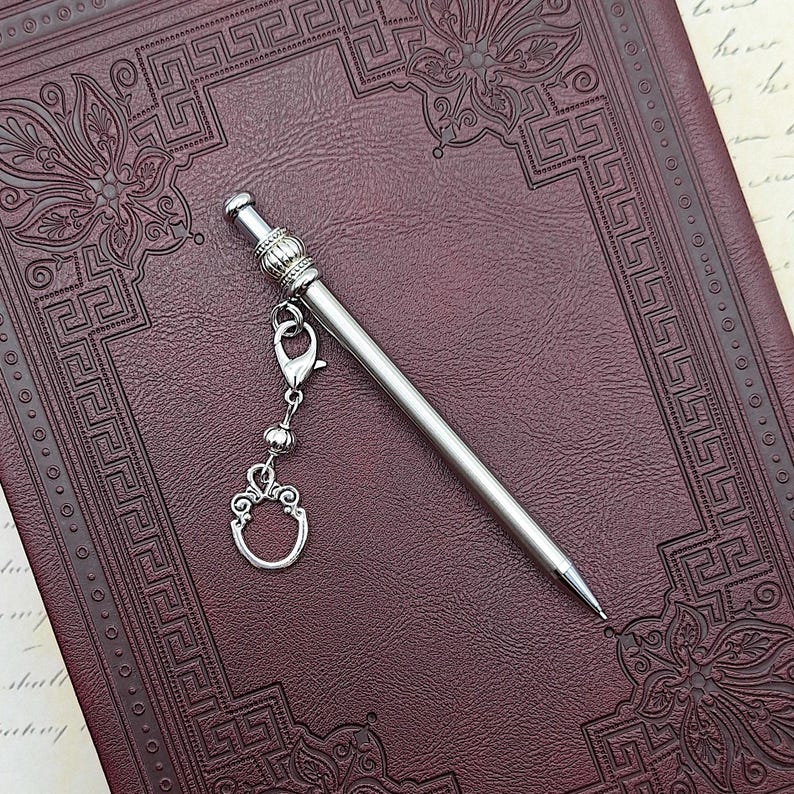 May include: A silver-toned metal mechanical pencil featuring a slender, smooth barrel and a decorative cap with a ribbed texture. A small silver charm in the shape of an ornate oval frame is attached to the pencil by a lobster clasp and chain.