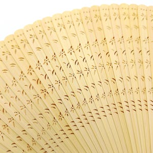 May include: A close-up of a light brown bamboo folding fan with a delicate floral pattern carved into the surface.
