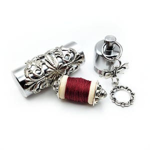 May include: A silver metal spool holder with a decorative floral design. The spool is filled with red thread and has a white wooden base. The holder is attached to a silver metal keychain with a small bow and a circular charm.