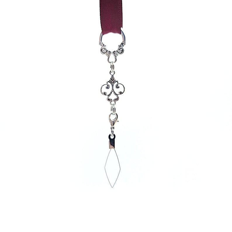 Pode incluir: Um marcador de livro de fita bord&ocirc; com um berloque decorativo prateado. O berloque apresenta um design ornamentado com um fecho de lagosta e um fio em forma de diamante. O marcador de livro est&aacute; sobre um fundo branco.