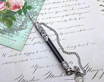 May include: A black pen with a silver-toned metal cap featuring intricate filigree patterns. The pen is attached to a silver-toned metal chain that connects to a small, circular ring charm with a floral design at the end of the chain.