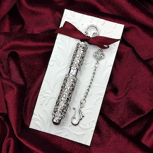 May include: A silver filigree pen with a chain and charm, displayed on a white background with a red ribbon.