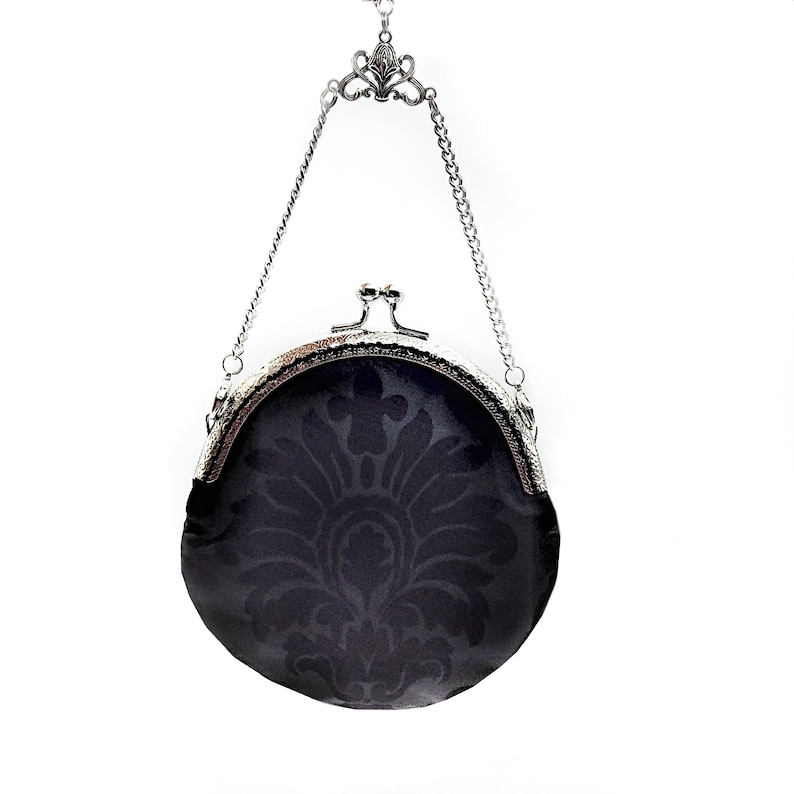 K&ouml;nnte beinhalten: Eine schwarze runde Handtasche mit einer silbernen Kette und einem Verschluss. Die Tasche hat ein Damastmuster.
