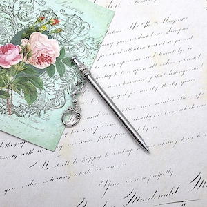 May include: A slim, silver-toned metal pen featuring a decorative cap with a small lobster clasp attachment. A dangling charm with an ornate, swirling metal design is connected to the pen by a short chain, resting against a surface.