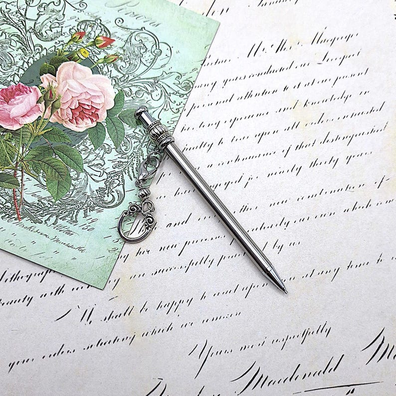 May include: A slim, silver-toned metal pen featuring a decorative cap with a small lobster clasp attachment. A dangling charm with an ornate, swirling metal design is connected to the pen by a short chain, resting against a surface.