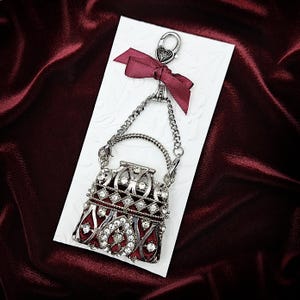 K&ouml;nnte beinhalten: Ein silberfarbener Handtaschenanh&auml;nger mit rotem Innenraum und besetzt mit klaren Strasssteinen. Der Anh&auml;nger h&auml;ngt an einer Kette und einer weinroten Schleife, die an einem silbernen Verschluss befestigt ist. Der Anh&auml;nger befindet sich auf einer wei&szlig;en Karte.