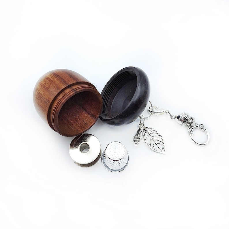 May include: A wooden acorn-shaped sewing kit with a black lid, a metal thimble, a metal needle threader, and a silver charm with a leaf design attached to a silver chain.