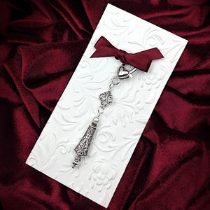 May include: A silver charm with a heart, a swirl design, and a pearlescent bead hangs from a silver chain. The charm is displayed on a white card with a burgundy ribbon.