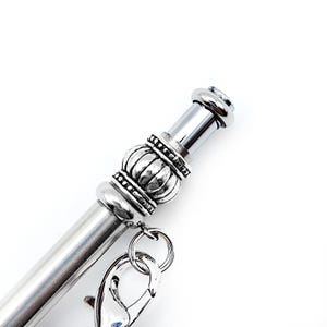 May include: A silver-toned metal pen featuring a decorative, ribbed cylindrical bead accent near the top. A small silver lobster claw clasp is attached to the bead by a jump ring, allowing for the addition of charms or other small accessories.