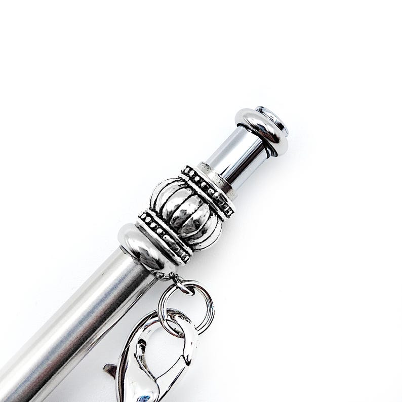 May include: A silver-toned metal pen featuring a decorative, ribbed cylindrical bead accent near the top. A small silver lobster claw clasp is attached to the bead by a jump ring, allowing for the addition of charms or other small accessories.