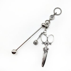 May include: Silver-toned decorative hair stick with a ball on one end and a charm with a button, a ball, and a pair of scissors on the other end.