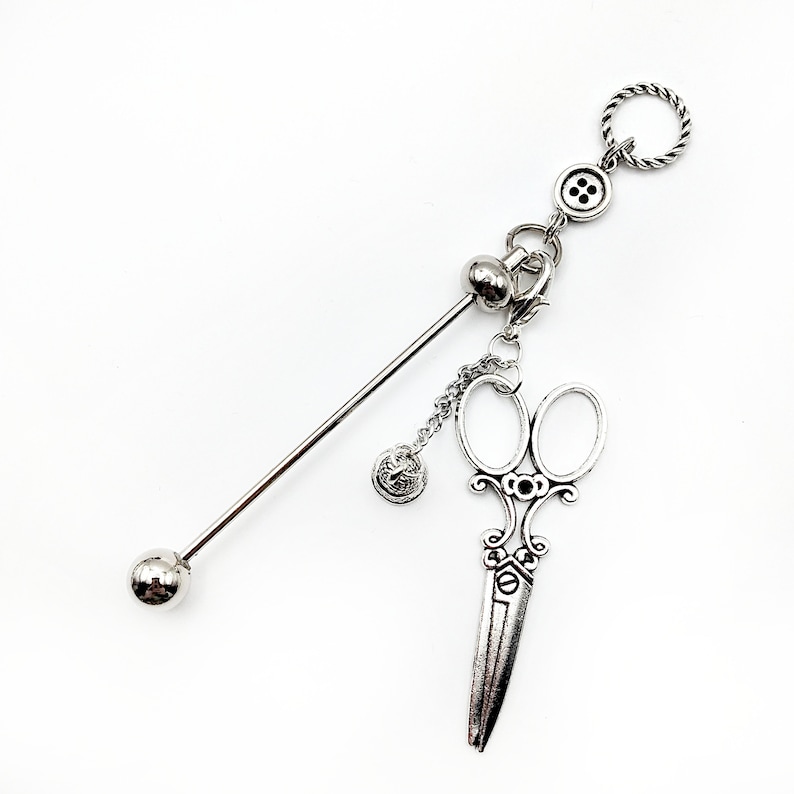 May include: Silver-toned decorative hair stick with a ball on one end and a charm with a button, a ball, and a pair of scissors on the other end.