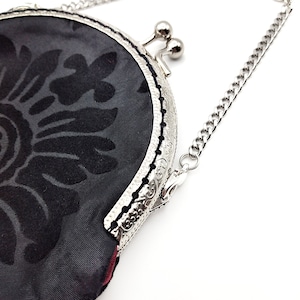 K&ouml;nnte beinhalten: Eine schwarze Stoffm&uuml;nzb&ouml;rse mit einer silbernen Kette und einem floralen Muster. Die B&ouml;rse hat einen silbernen Verschluss und einen silbernen Besatz.