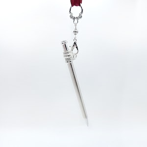 May include: A silver-toned metal stylus pen featuring a decorative filigree cap and a ribbed barrel design. The pen is suspended from a dark red ribbon by a silver-colored lobster clasp and a small metal bead connector.