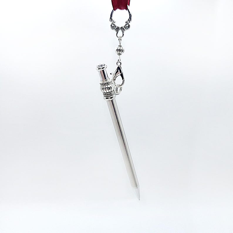 May include: A silver-toned metal stylus pen featuring a decorative filigree cap and a ribbed barrel design. The pen is suspended from a dark red ribbon by a silver-colored lobster clasp and a small metal bead connector.