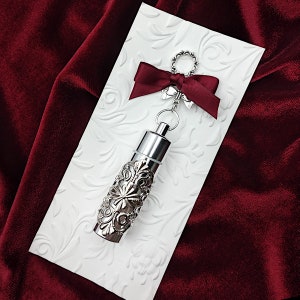 K&ouml;nnte beinhalten: Ein silbernes Medaillon mit einem verzierten Design, das an einer silbernen Kette mit einer roten Schleife befestigt ist. Das Medaillon befindet sich auf einem wei&szlig;en Hintergrund mit einem floralen Muster.