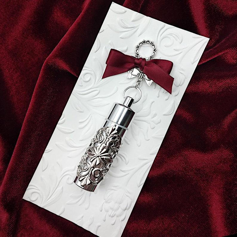 K&ouml;nnte beinhalten: Ein silbernes Medaillon mit einem verzierten Design, das an einer silbernen Kette mit einer roten Schleife befestigt ist. Das Medaillon befindet sich auf einem wei&szlig;en Hintergrund mit einem floralen Muster.