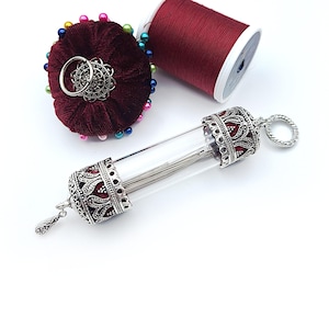 May include: A burgundy velvet pin cushion with colorful pins, a spool of red thread, and a glass needle case with ornate silver-colored metal ends. The needle case contains several needles.