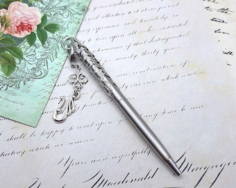 May include: A silver-toned metal ballpoint pen featuring an intricate embossed floral pattern on the upper barrel and a dangling silver charm shaped like a decorative fleur-de-lis symbol attached to the clip by a small metal link.