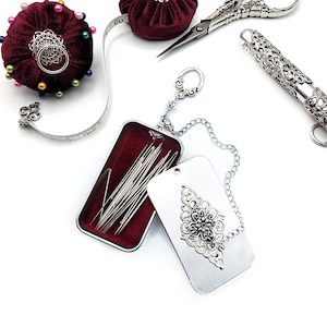 May include: A silver metal needle case with a chain and a decorative charm. The case is open and filled with needles. A red pincushion with colorful beads is in the background.