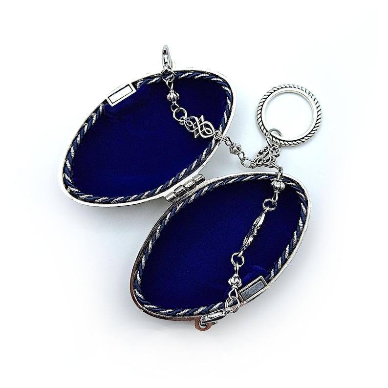 May include: An open, silver-toned oval locket with a dark blue velvet interior. The locket is connected to a key ring by a delicate chain. The edges are trimmed with a twisted rope design. The locket is on a white background.
