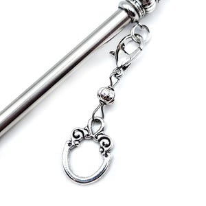 May include: A silver-toned metal hair stick featuring a smooth, cylindrical shaft and a decorative top. A silver-colored charm dangles from a lobster claw clasp, consisting of a small round bead and an oval-shaped frame with scrollwork details.