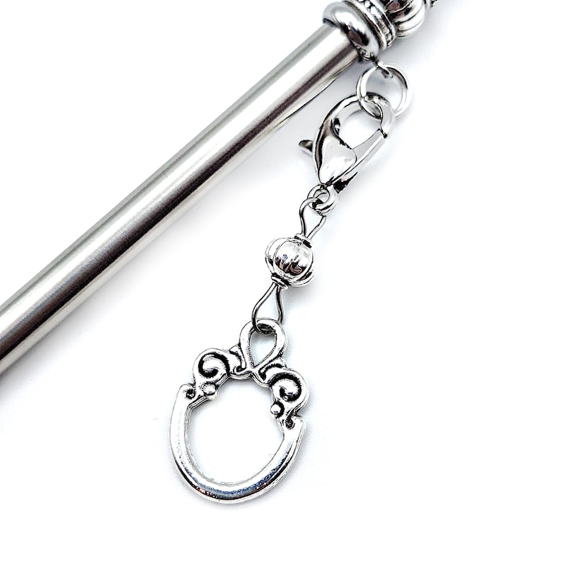 May include: A silver-toned metal hair stick featuring a smooth, cylindrical shaft and a decorative top. A silver-colored charm dangles from a lobster claw clasp, consisting of a small round bead and an oval-shaped frame with scrollwork details.