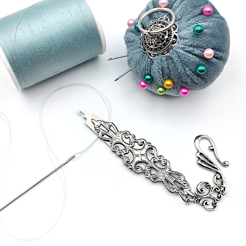 May include: A silver needle threader with an ornate design and a hook on the end. A spool of light blue thread, a needle, and a pincushion with colorful beads are also in the image.