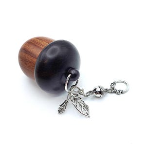 May include: A wooden acorn-shaped box with a silver chain and charms, including a leaf and an acorn. The box is made of dark wood and has a light brown top.