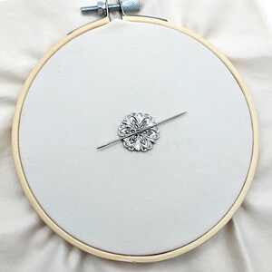 May include: A silver embroidery hoop with a decorative metal flower design and a needle threaded through the center.