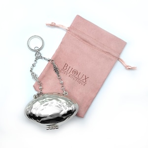 May include: A silver-colored, hammered metal purse with a chain and ring handle, next to a pink drawstring bag. The bag has the text "BIJOUX GIFT BOUTIQUE" printed on it. The purse has a hinged bottom.