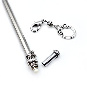May include: A silver-toned metal hair stick featuring a decorative, ribbed, and beaded end cap with a small white tip. A separate silver-toned metal charm with a lobster clasp and a circular, ornate filigree pendant is positioned nearby.