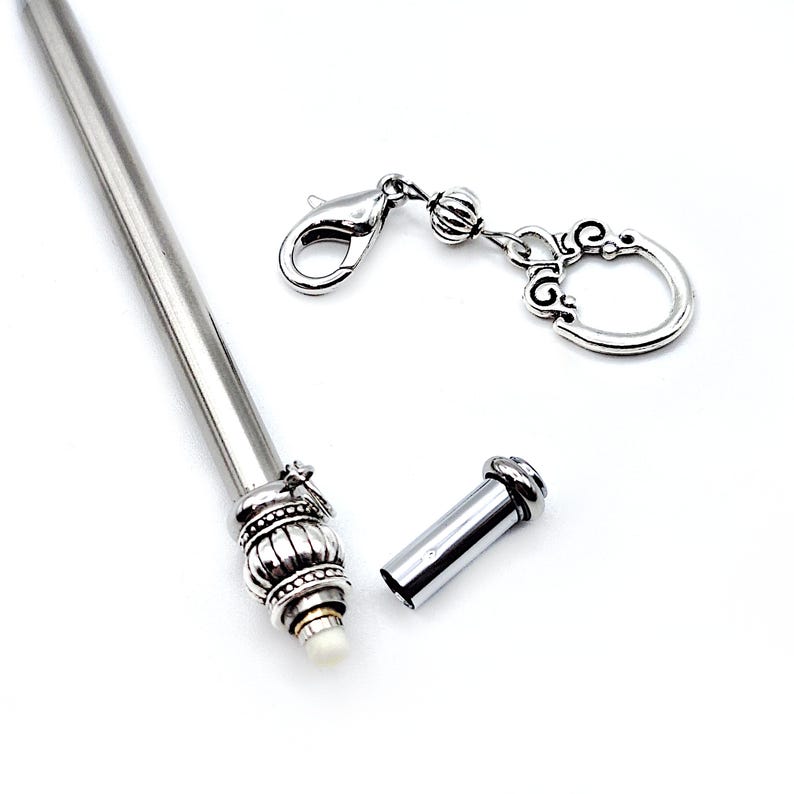 May include: A silver-toned metal hair stick featuring a decorative, ribbed, and beaded end cap with a small white tip. A separate silver-toned metal charm with a lobster clasp and a circular, ornate filigree pendant is positioned nearby.