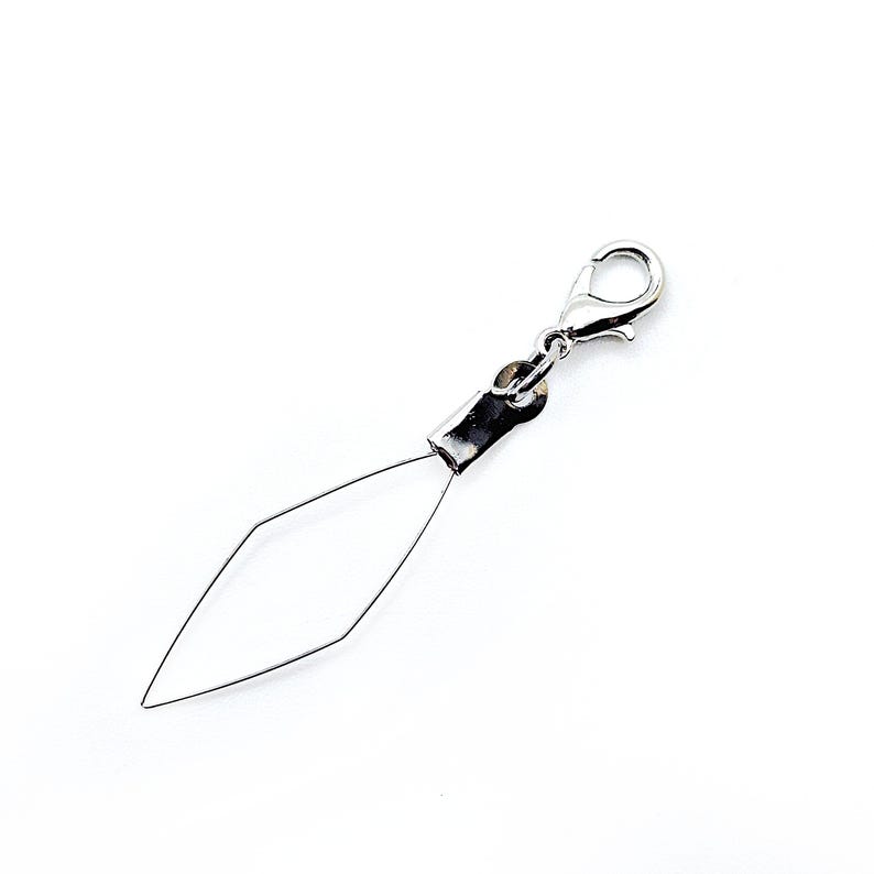 Pode incluir: Um passador de agulha prateado com um fecho de lagosta. O passador apresenta um la&ccedil;o de arame em forma de diamante preso a uma manga e fecho de metal, projetado para facilitar a passagem da agulha. O fundo &eacute; branco liso.