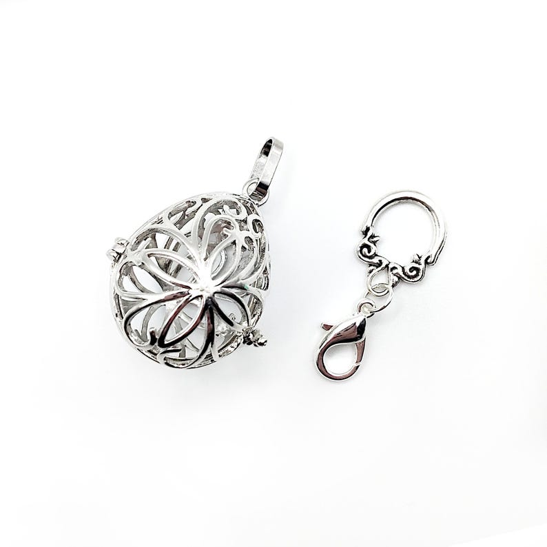 May include: A silver charm with a delicate, intricate design. The charm is shaped like a teardrop and has a small loop at the top for attaching to a necklace or bracelet. The charm is made of metal and has a shiny finish.