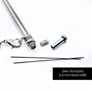 May include: A silver mechanical pencil with a decorative ribbed end, a lobster clasp attachment, a small eraser, a metal cap, and two thin black leads. The text on the image states: Uses standard 0.5 mm lead refills.